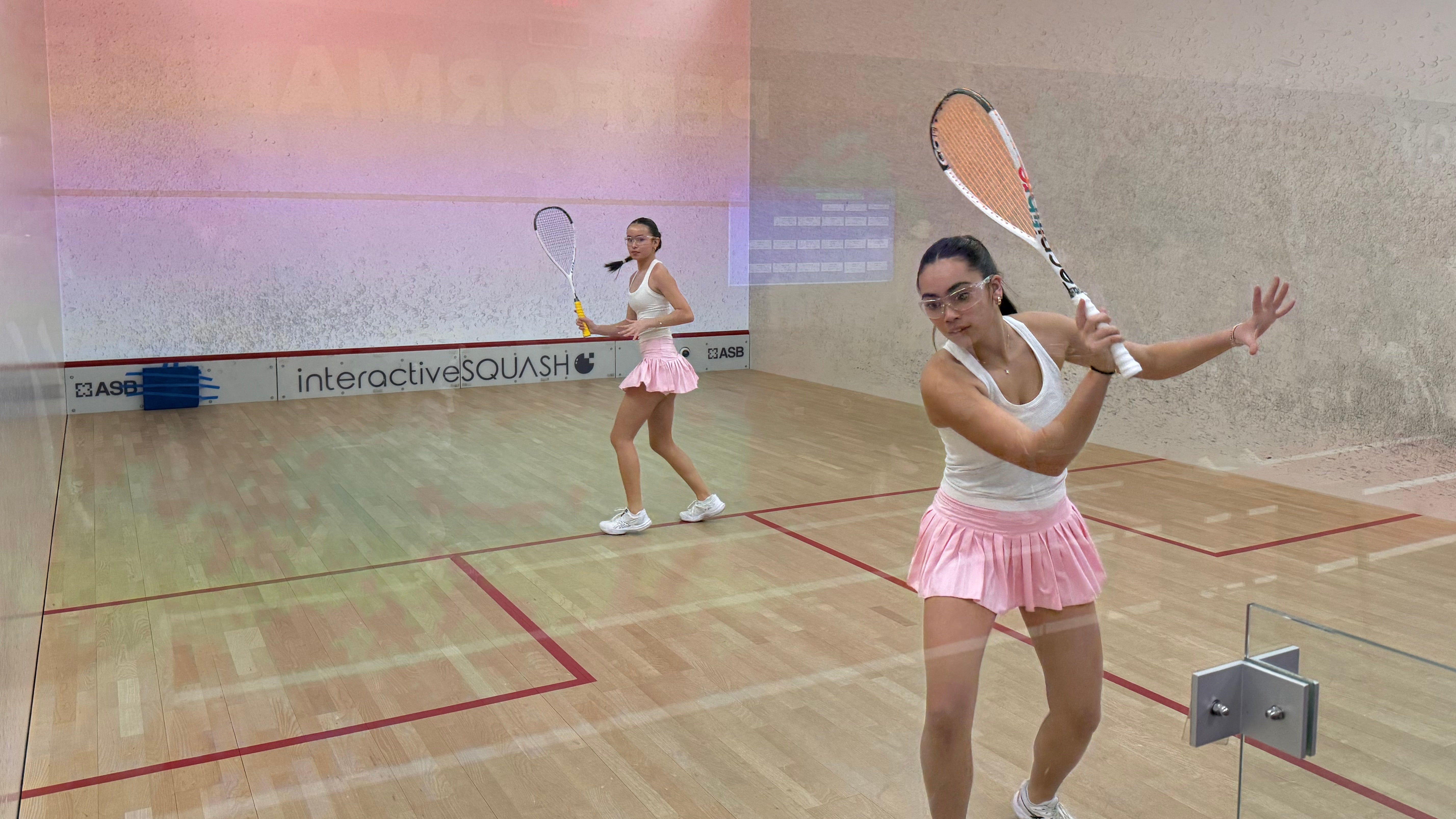 Two women playing squash in a indoor court with 'interactiveSQUASH' branding.