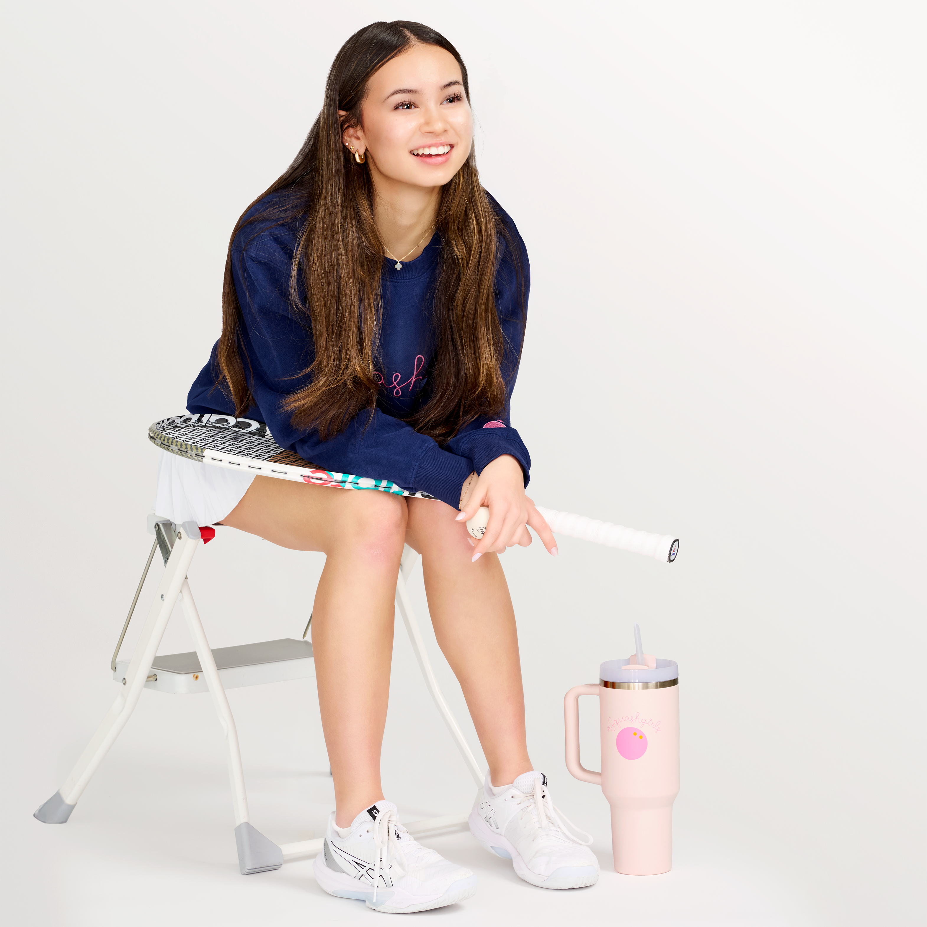 Girl sitting on a stool with a tennis racket, wearing a navy blue crew neck sweatshirt with pink text, on a white background.